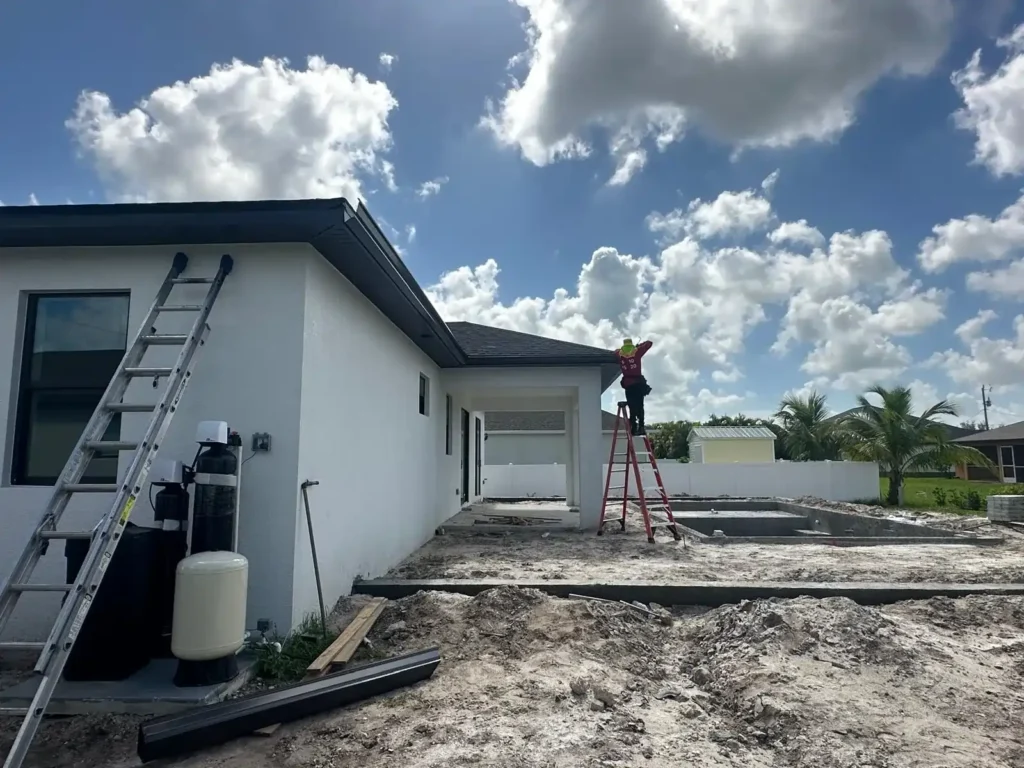 A construction worker installs seamless gutters on a white single-story house under a clear blue sky.