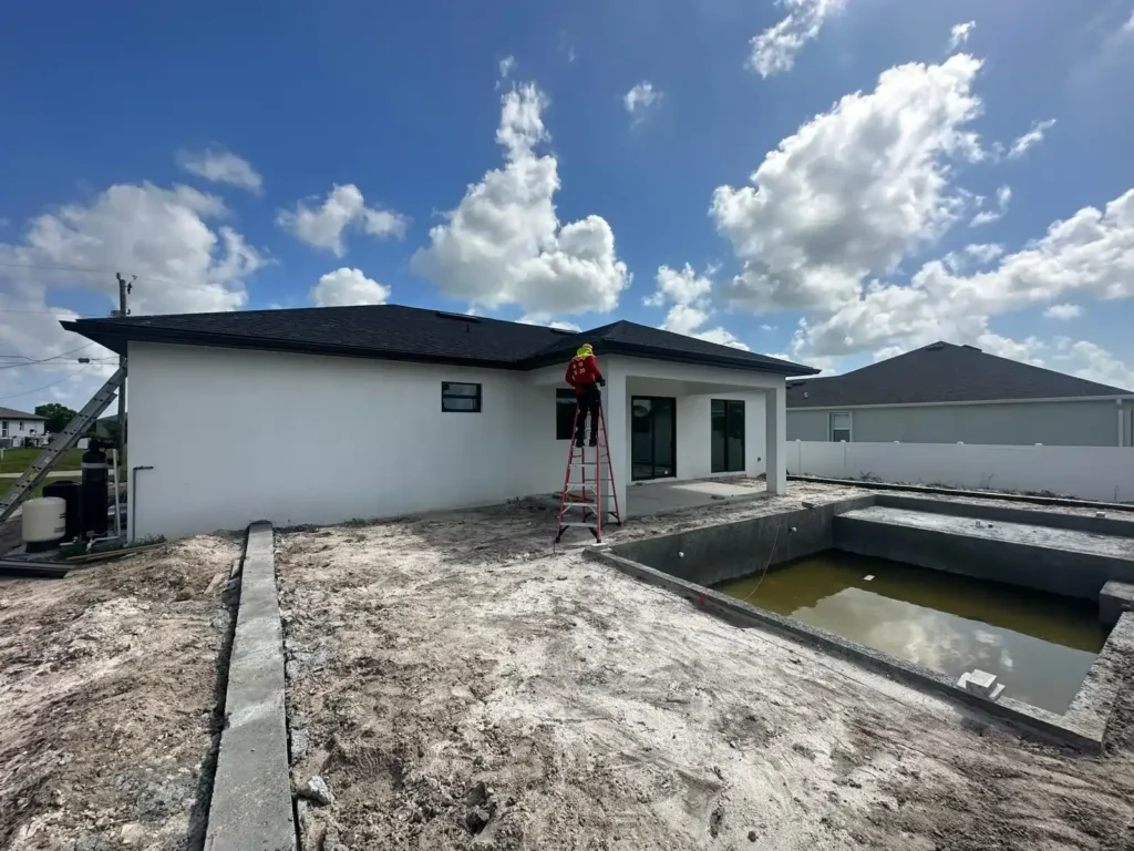 A construction worker stands on a red ladder installing seamless gutters on a white house with an in-progress pool in the backyard.